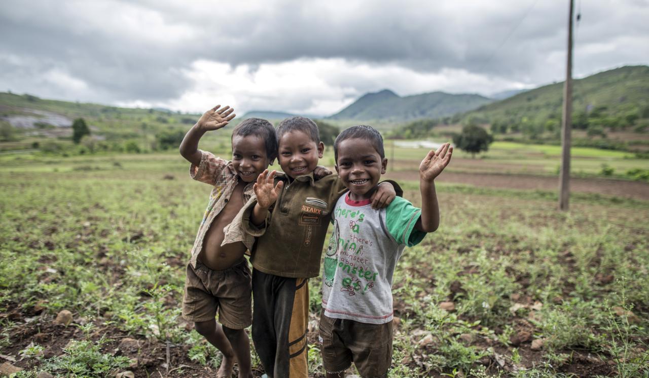 Three smiling boys in a field wave at the camera. 