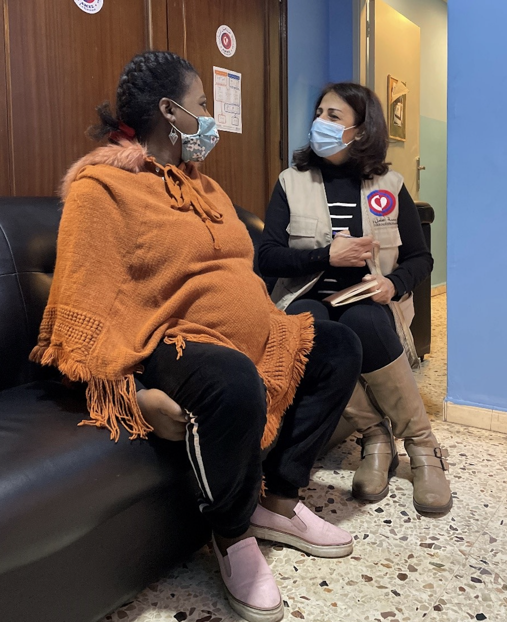 Dos mujeres, una con jersey naranja y otra con camisa negra y chaleco terracota, se sientan en un sofá a hablar entre ellas. 
