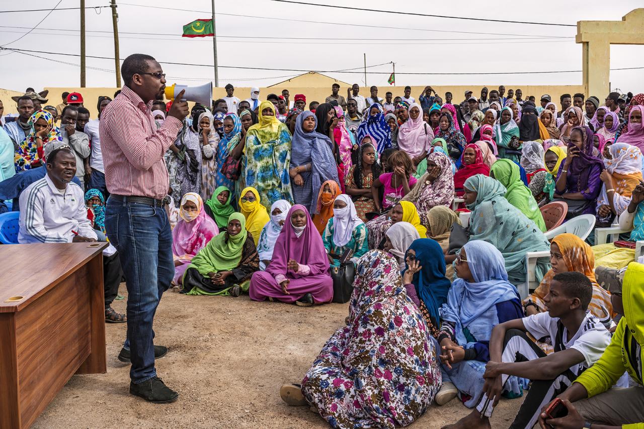Many people in multi-coloured garb sit around a man speaking at a desk. 