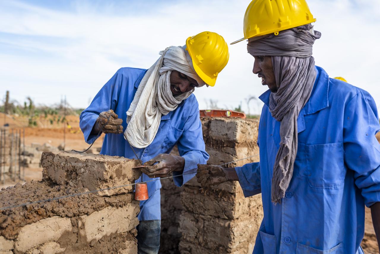 Two people in hard hats examine bricks as part of a construction site. 