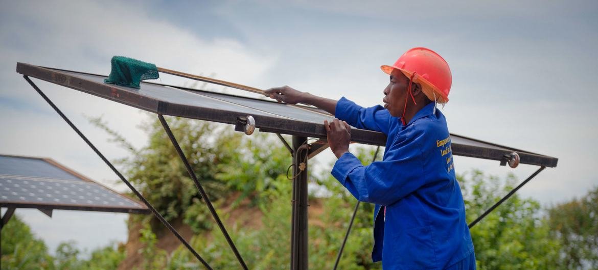 Un homme portant un bleu de travail et un casque de protection rouge nettoie un panneau solaire en extérieur.