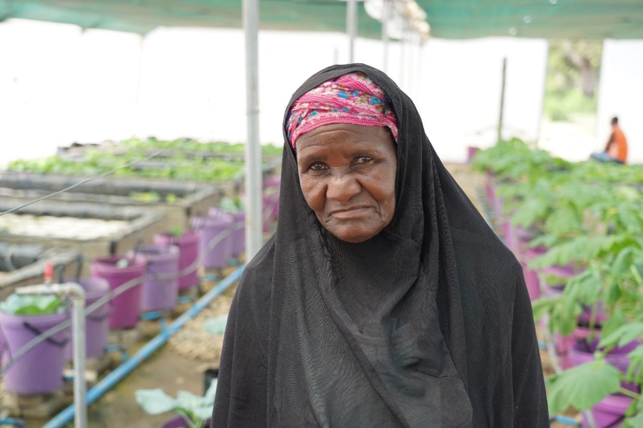 A woman in a black veil looks at the camera, and stands in between a row of plants in a greenhouse.