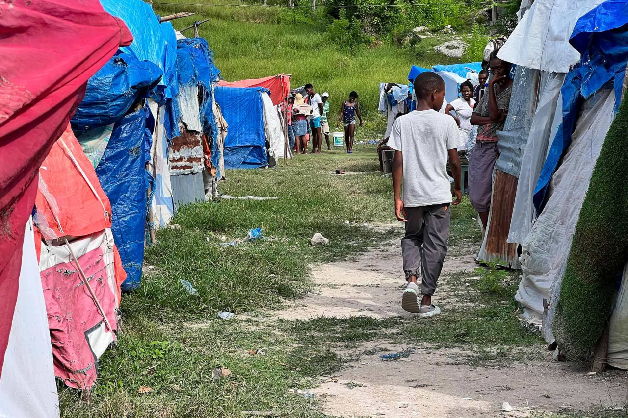 Un joven es fotografiado de espaldas caminando por un callejón bordeado por dos hileras de tiendas de campaña hechas con lonas de diferentes colores.