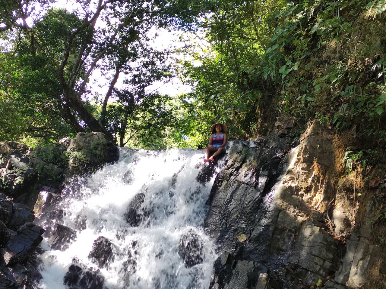 Una mujer sentada en medio del borde de una pequeña cascada en un área verde.