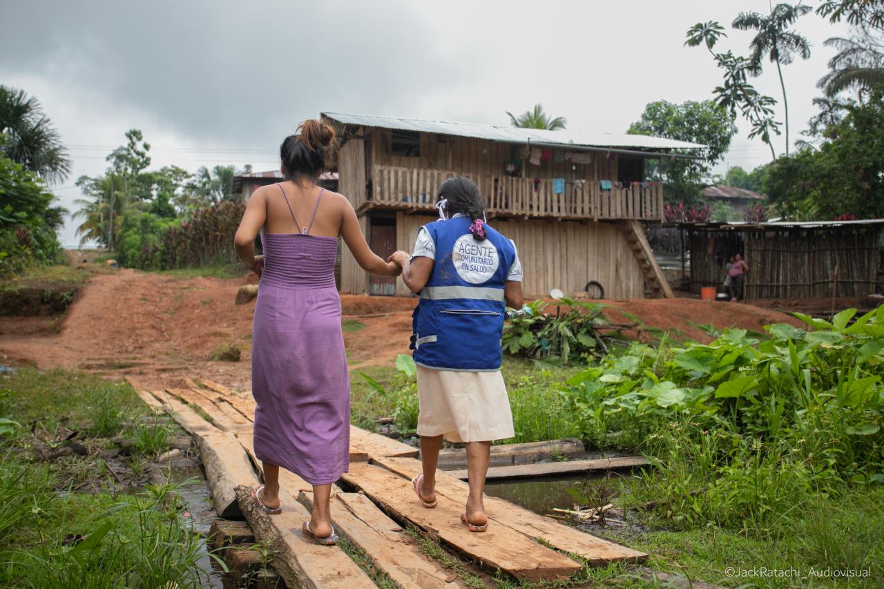 Dos mujeres caminando sobre unas tablas de madera en un espacio abierto lleno de vegetación tropical, y se dirigen hacia una edificación de dos plantas.