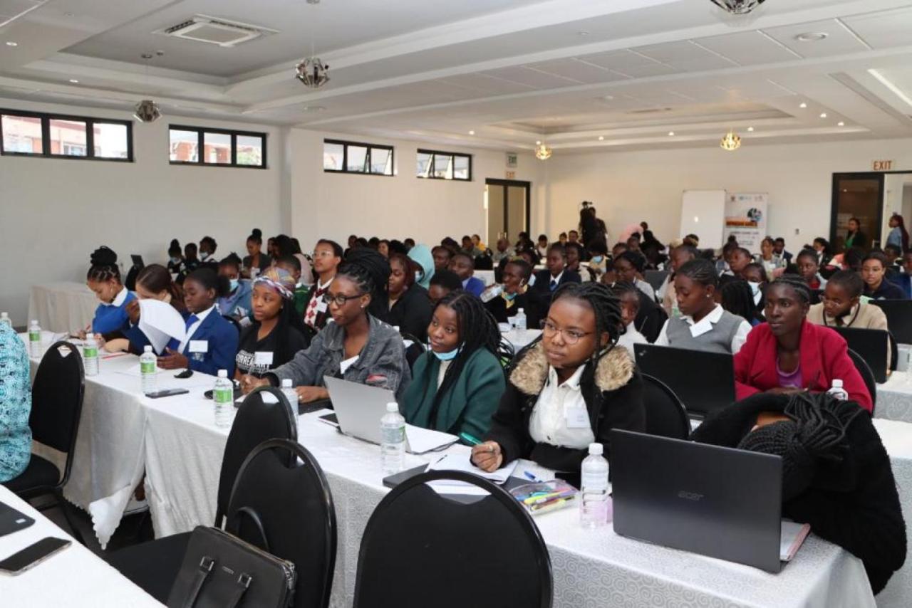 Young girls are sitting in a room, in front of laptops placed on large tables covered with white tablecloths, carefully listening to their trainer in a coding course.