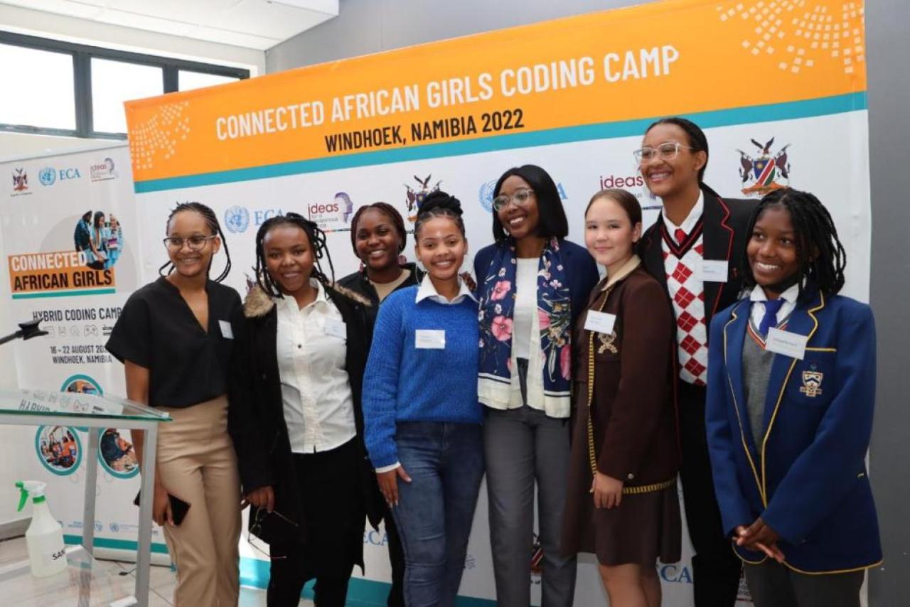 A group of girls stand side by side with an older woman in front of a banner that reads "Connected African Girls Coding Camp" and smile at the camera.