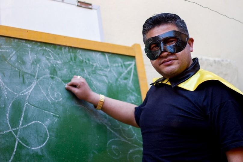 El súper profe boliviano disfrazado con una tiza en la mano frente a una pizarra.
