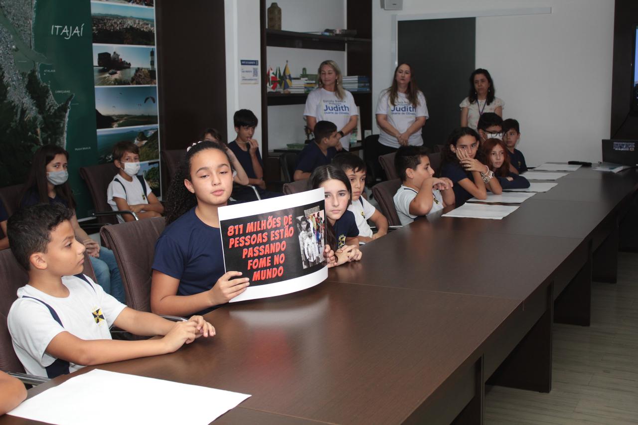 A group of children are sitting in a meeting room with their teacher at a special recognition event in the Mayor's office at  Itajai's City Hall.