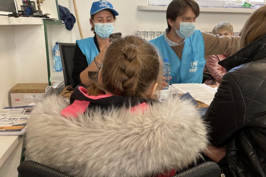 Un homme et une femme tous deux vêtus d'un gilet bleu et portant un masque de protection respiratoire s'entretiennent avec une femme et une fillette photographiées de dos et portant des vêtements chauds.