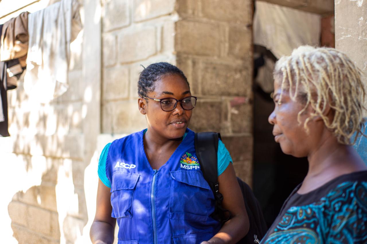 Close-up of a woman wearing glasses talking with another woman with bleached hair outside a house.