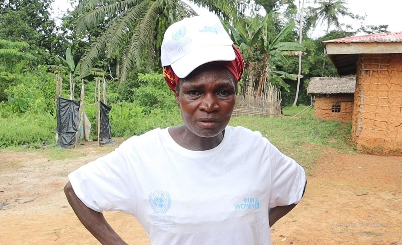 Portrait d'une femme libérienne portant un t-shirt blanc et une casquette blanche d'ONU Femmes se tenant debout devant une habitation en terre cuite entourée de végétation. 