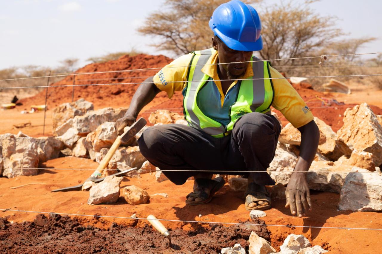 Man in a green vest and a blue hard hat kneels on the ground with tools