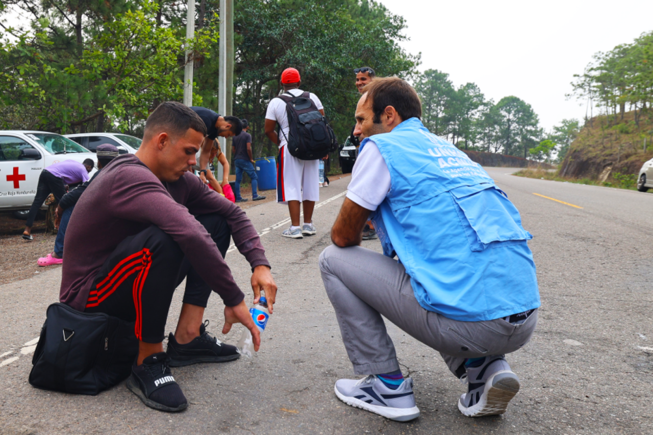 A man in a blue vest kneels on the ground next to another man crouched over, in a red shirt