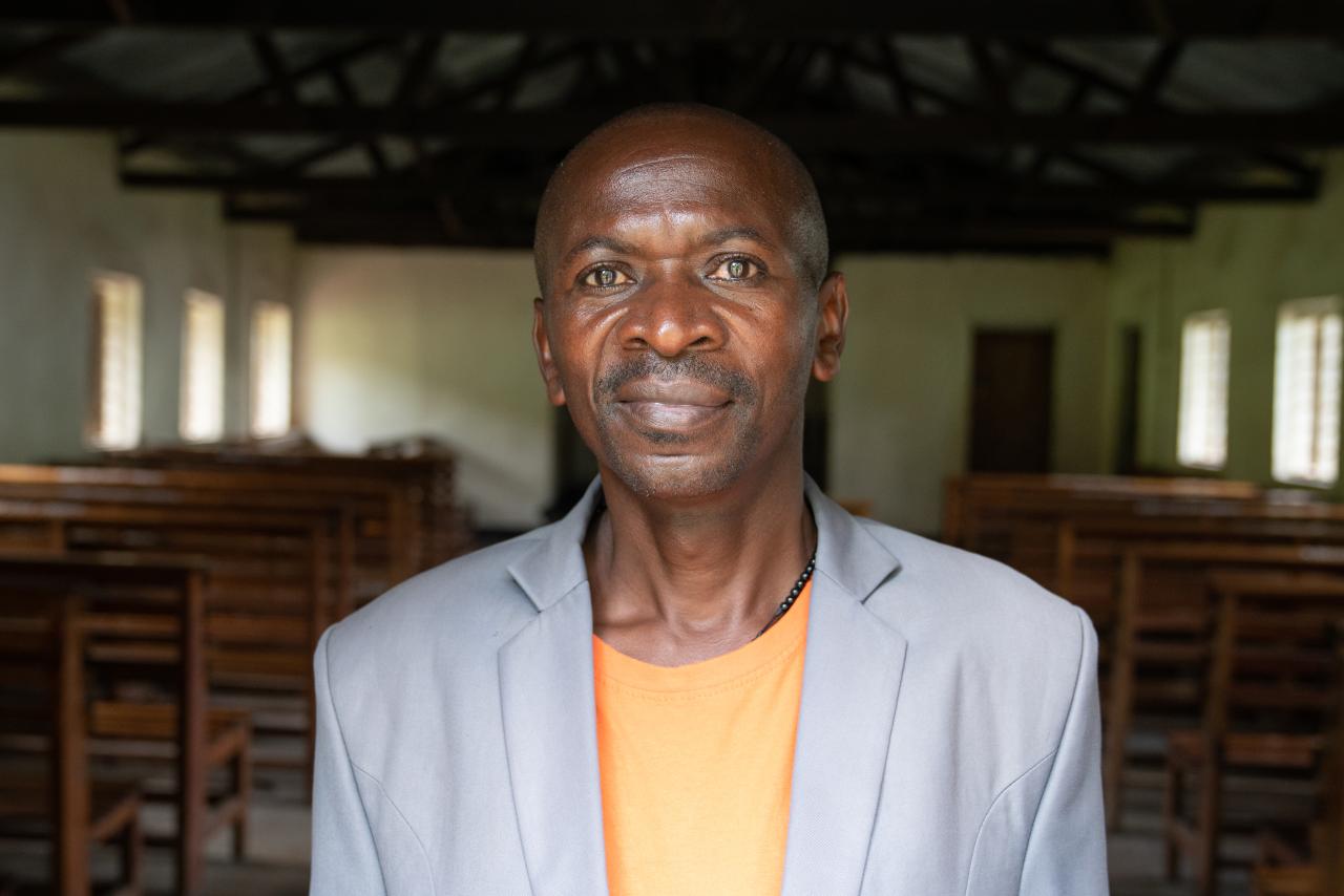 man in grey suit and orange top stands close to the camera, with a church interior behind him
