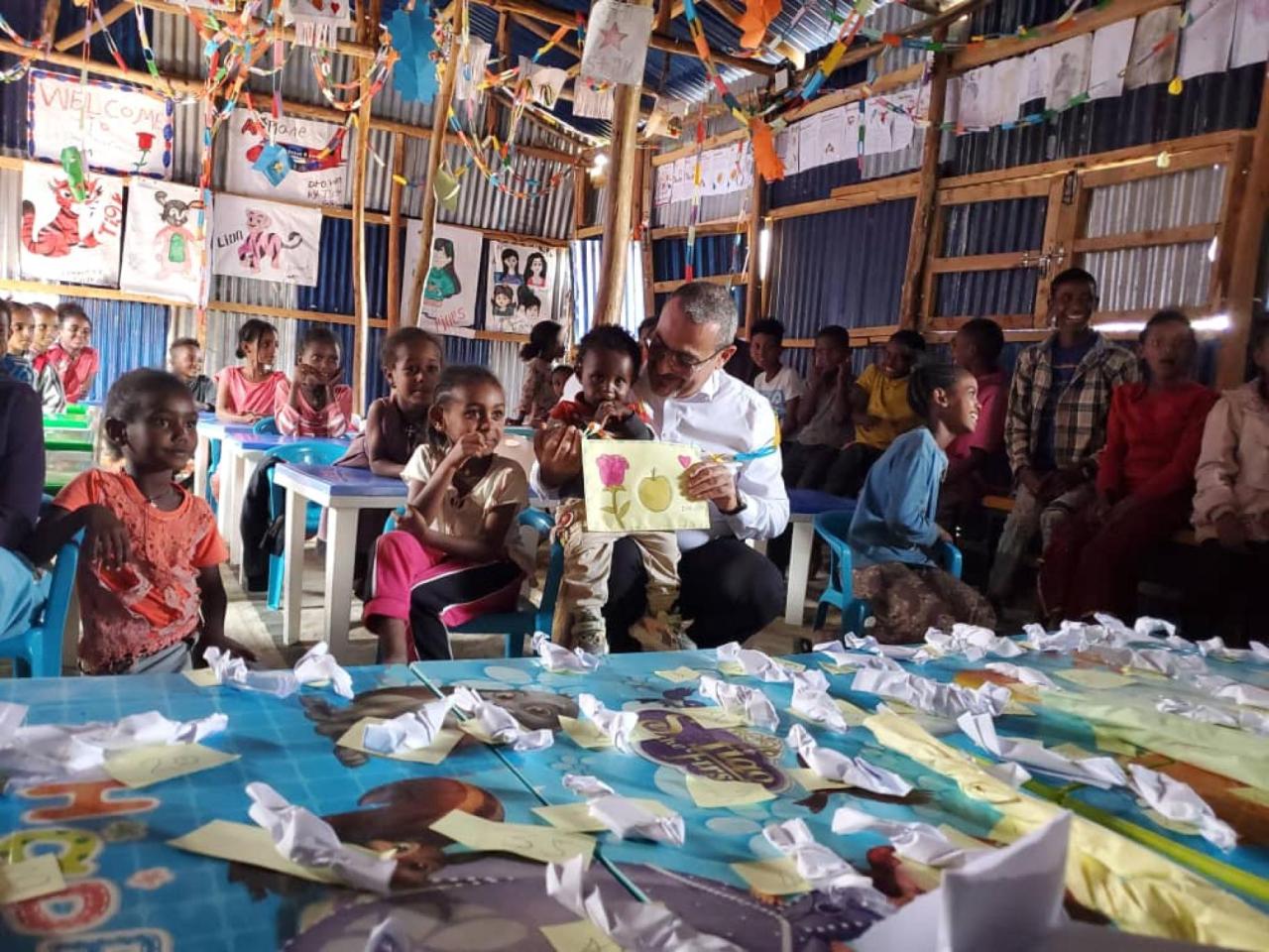 A group of children in a classroom where in the middle an older man sits on a small chair reading a book to them.
