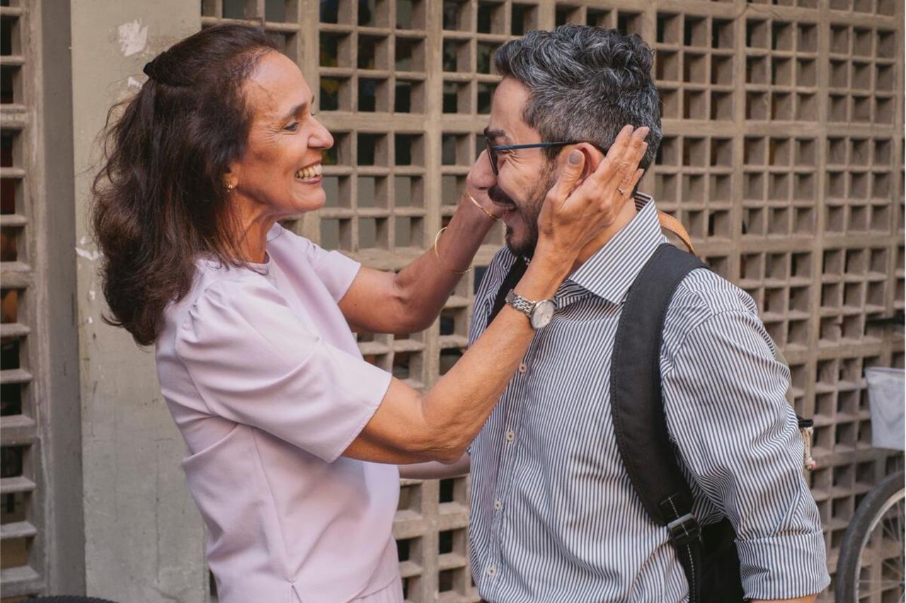 A woman in a pink shirt smiles and holds the face of a man in a grey shirt and glasses.