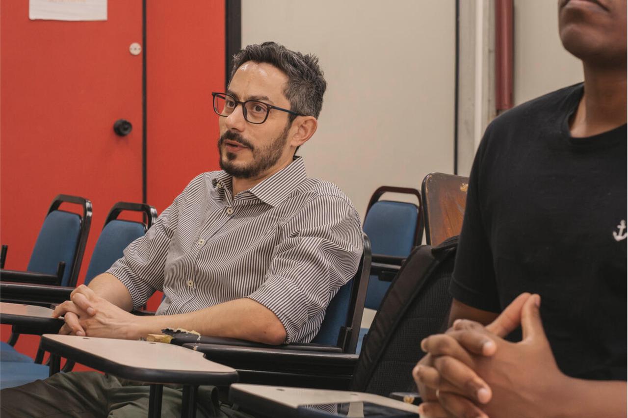 A man in a grey shirt and glasses sits at a desk in a classroom with bright orange walls behind him.
