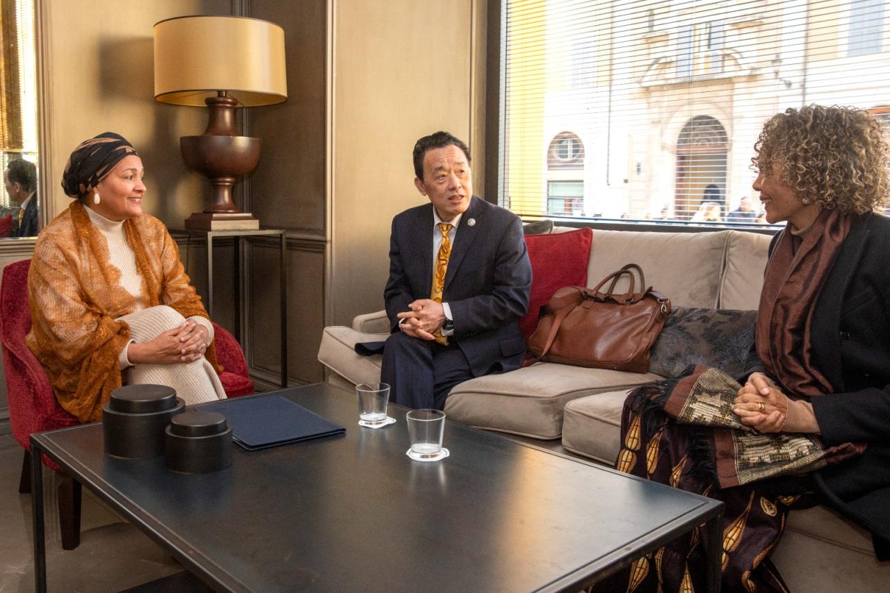 A woman in a lacey, brick-brown shawl and stylish headwrap (left) holds a meeting with two UN officials, all seated on couches in an informal setting.