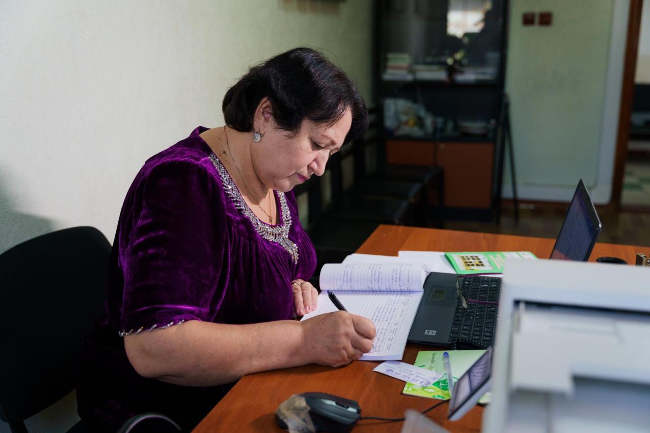 A woman in a purple dress sits at a desk writing in a notebook, papers strewn all around her.