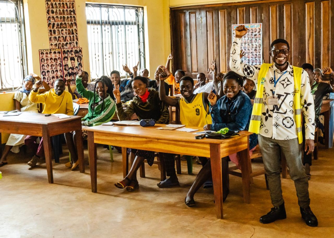 A man in a yellow and white shirt raises his hand in joy as he stands in front of a classroom full of students at desks who are also doing the same