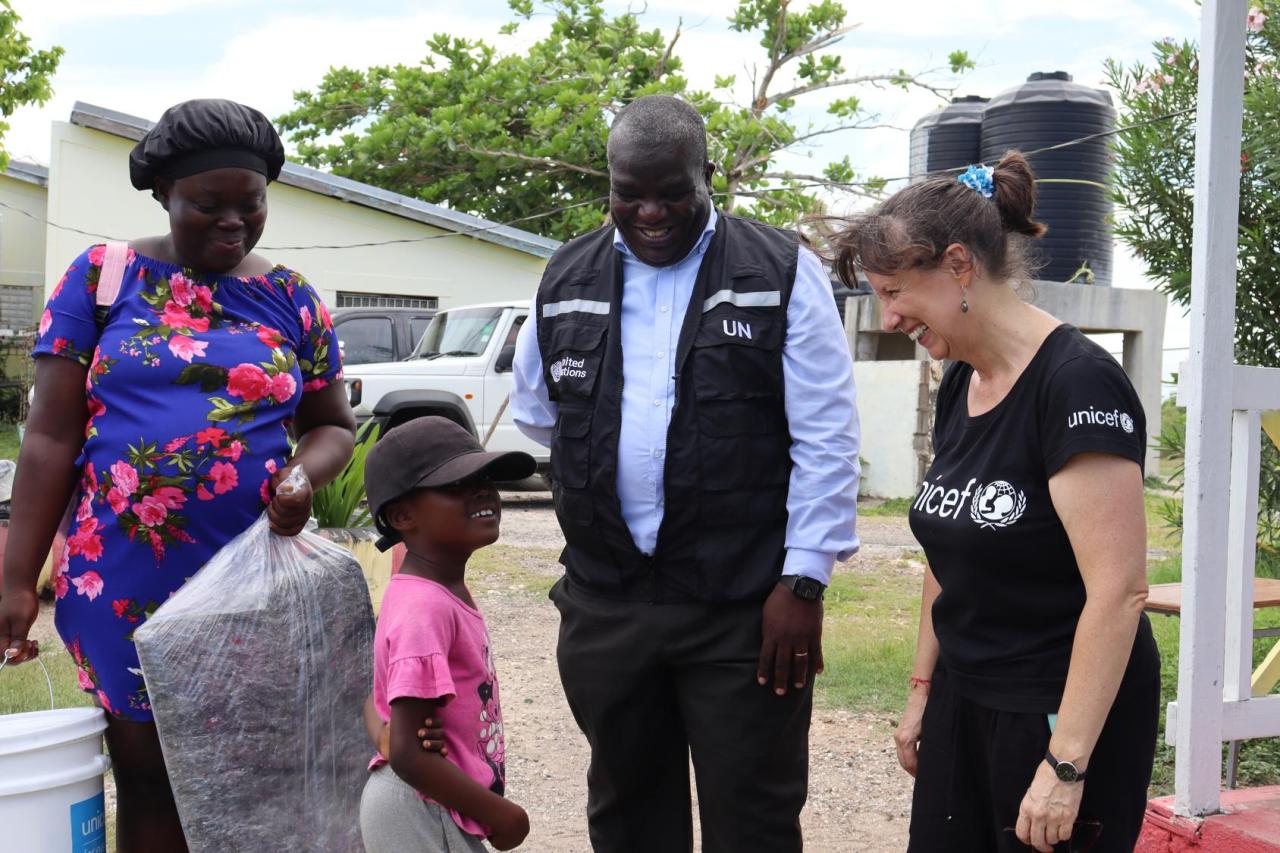 A man and a woman watch a little child carrying a bag