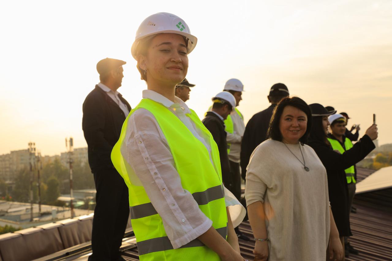 A woman in a green vest and hard hat smiles to the camera as other look on