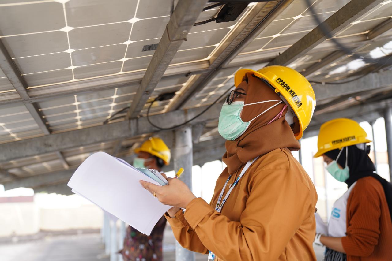 A woman in orange clothes and a hard hat stands under a solar panel