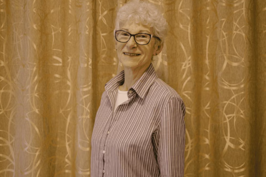 A pensioner in glasses and a striped shirt stands and smiles to the camera.