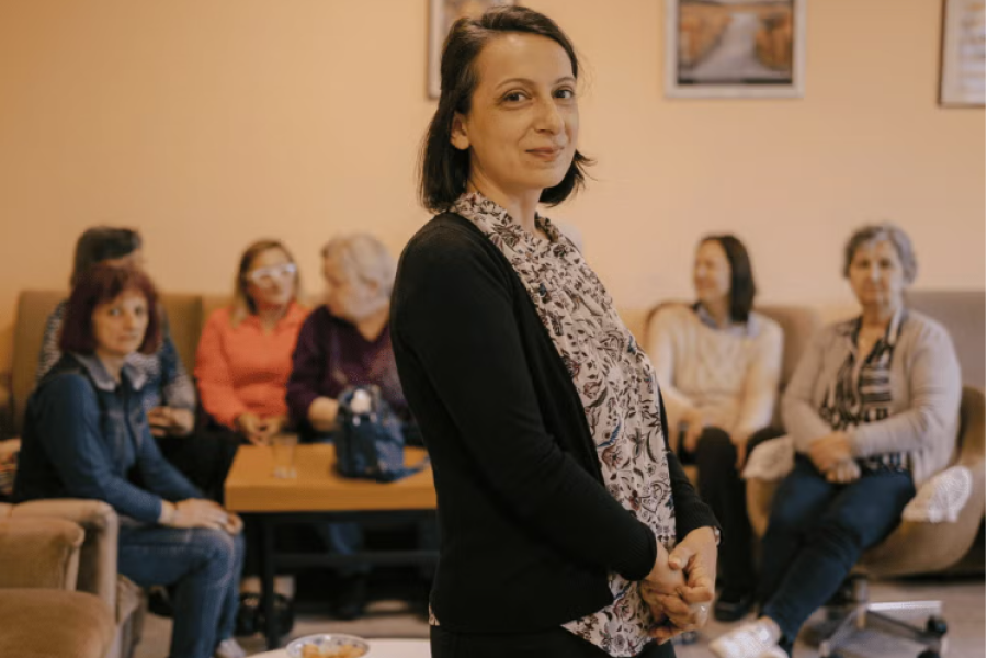 A pensioner stands and smiles to the camera, in the background a group of other pensioners are chatting amongst themselves.