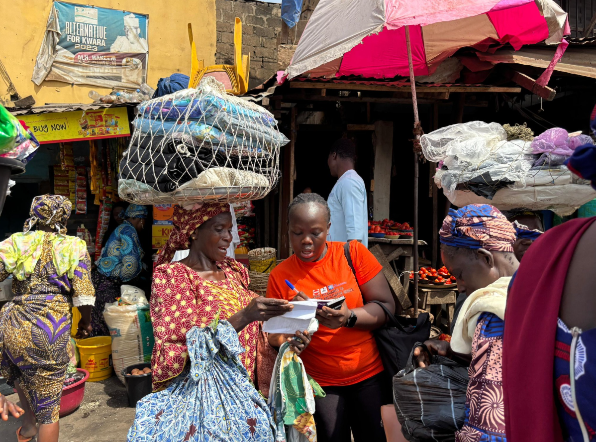 A Nigerian female volunteer stands in a busy marketplace and writes something down while talking to another woman.