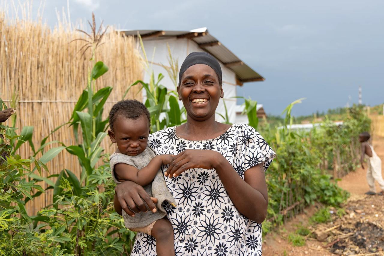 A mother in Burundi holds her baby and smiles in front of maize crops.