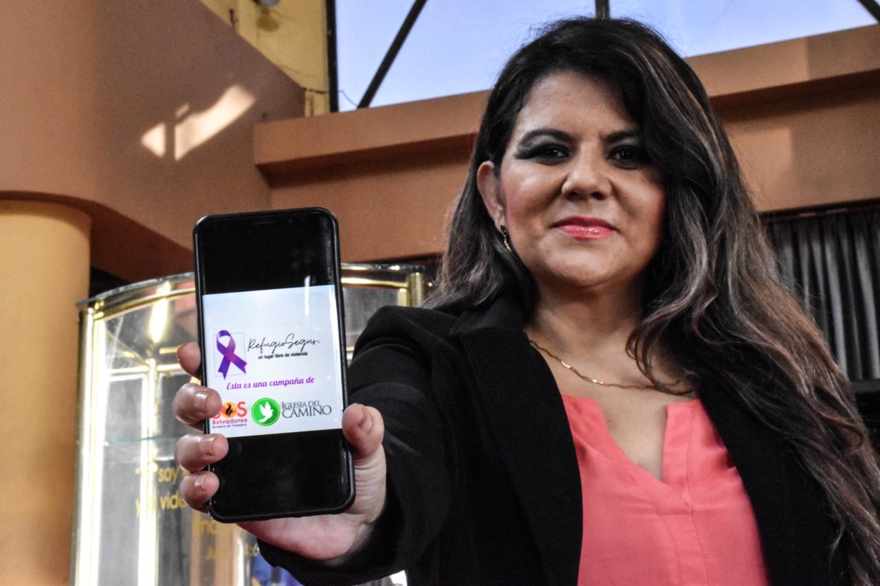 Una mujer que mira a la cámara sostiene un dispositivo móvil en cuya pantalla aparece un lazo morado y las palabras "Refugio Seguro". 