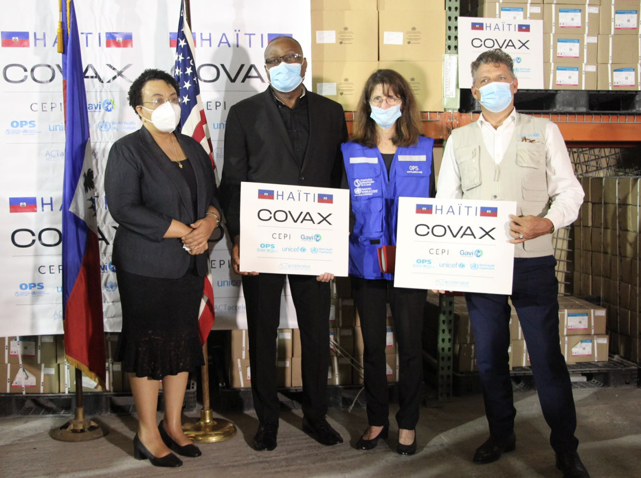 Cuatro personas sostienen carteles de COVAX delante de las banderas de Haití y Estados Unidos.