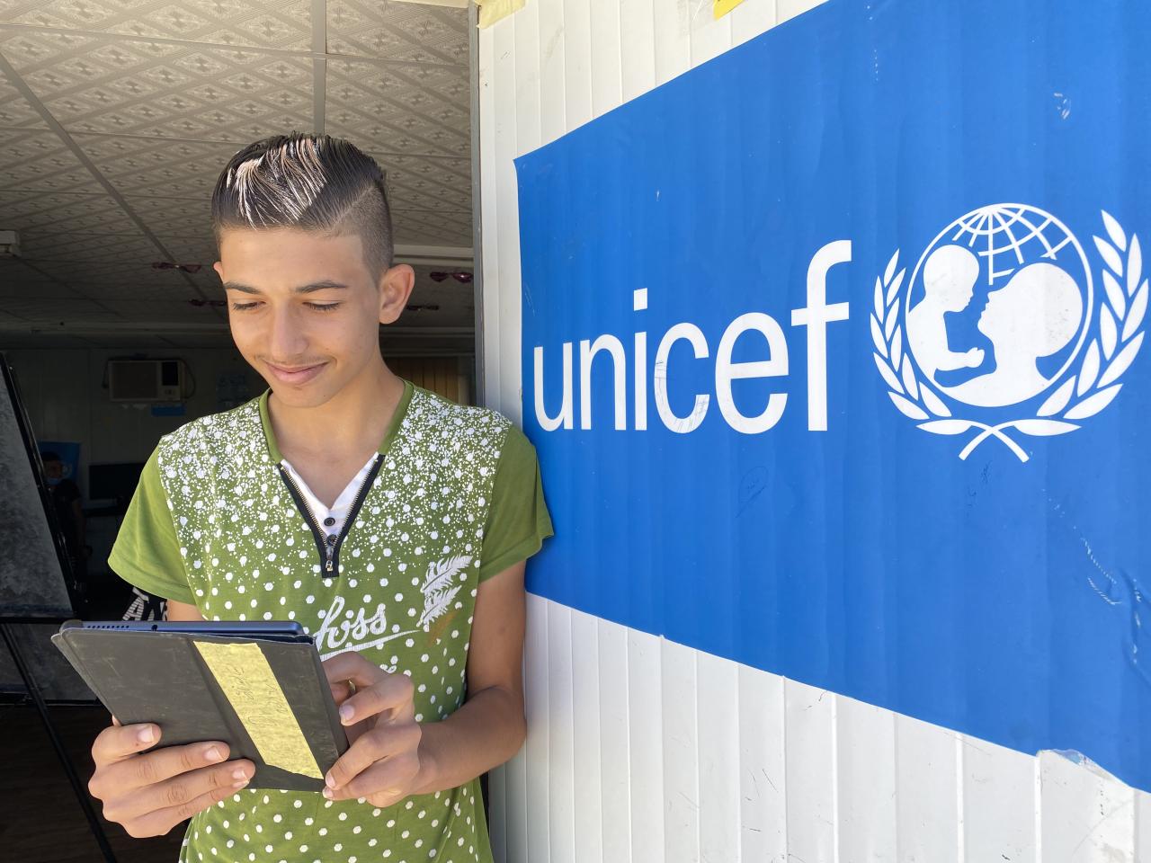 A boy smiles while he looks down at his tablet near a blue UNICEF sign. 