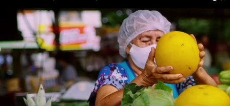 A woman in a hairnet and a white face mask holds a large yellow fruit in her hands. 