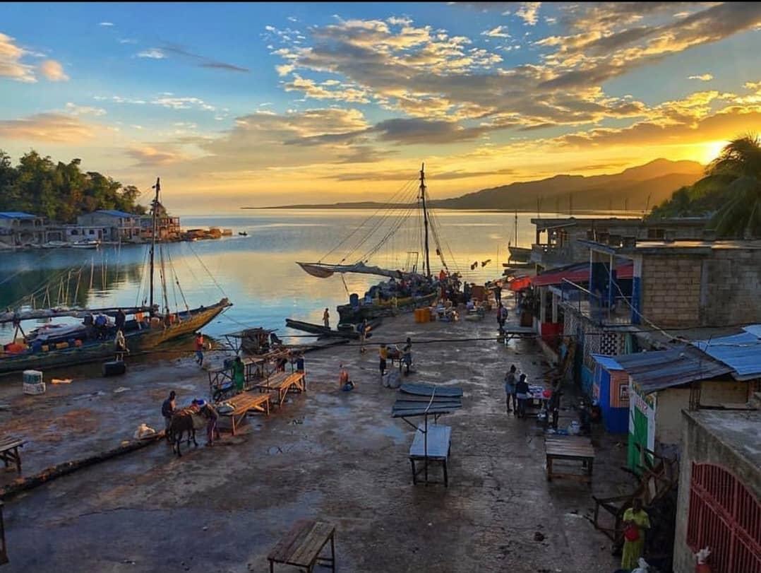 Several stores lined up near the ocean and people walking around the market during sunset. 