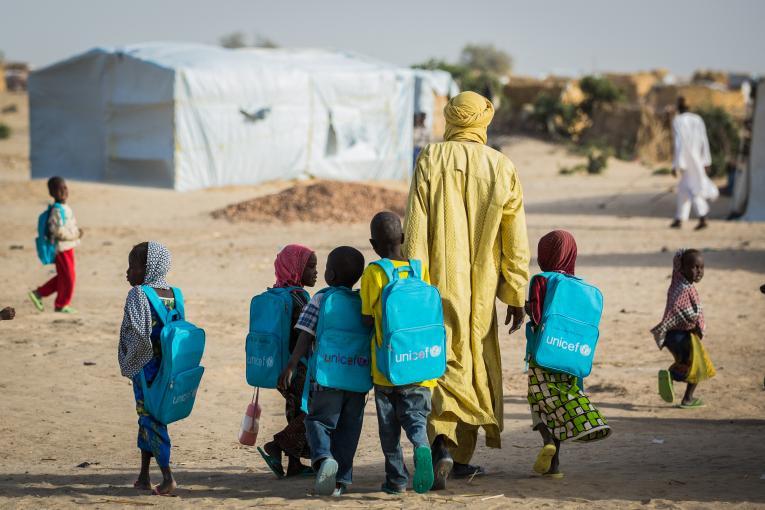 Un homme vêtu d'un habit traditionnel et d'un turban jaunes marche avec de jeunes enfants portant des sacs à dos bleus de l'UNICEF sur le dos. Dos à la caméra, le groupe se dirige vers une grande tente qui abrite une école.
