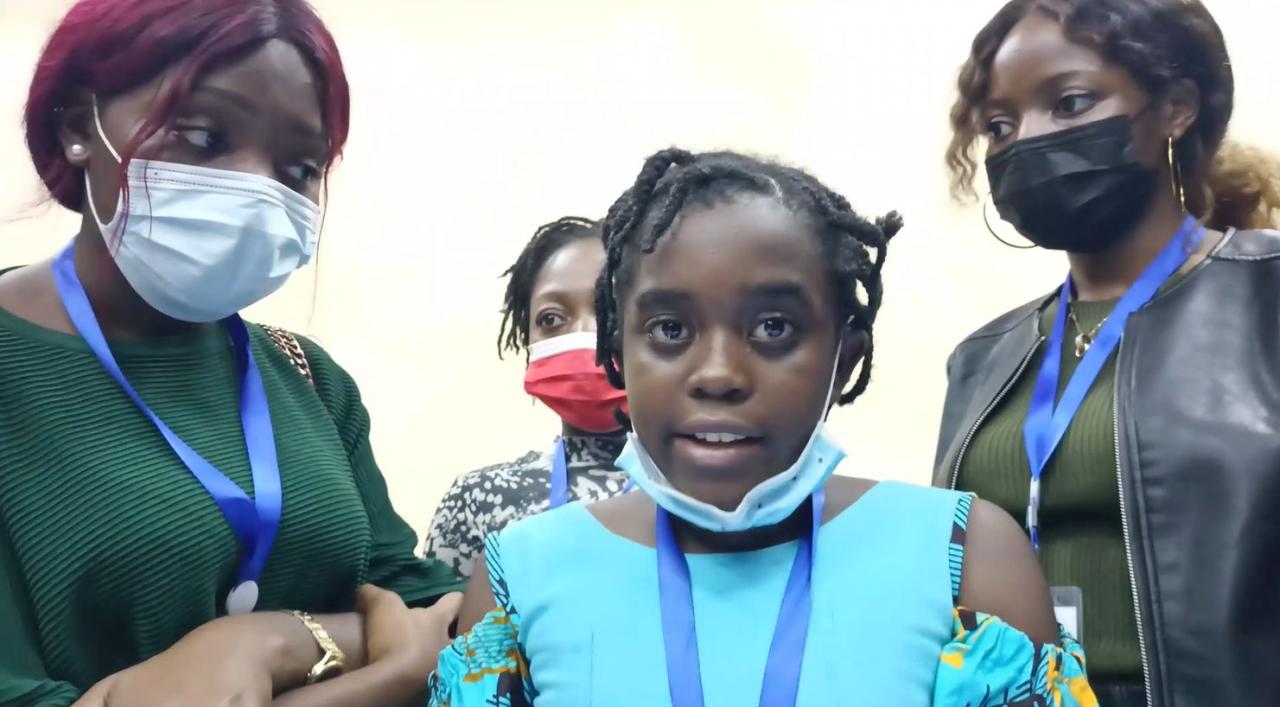 A young girl in a light blue shirt looks directly at the camera and is surrounded by three other women. 