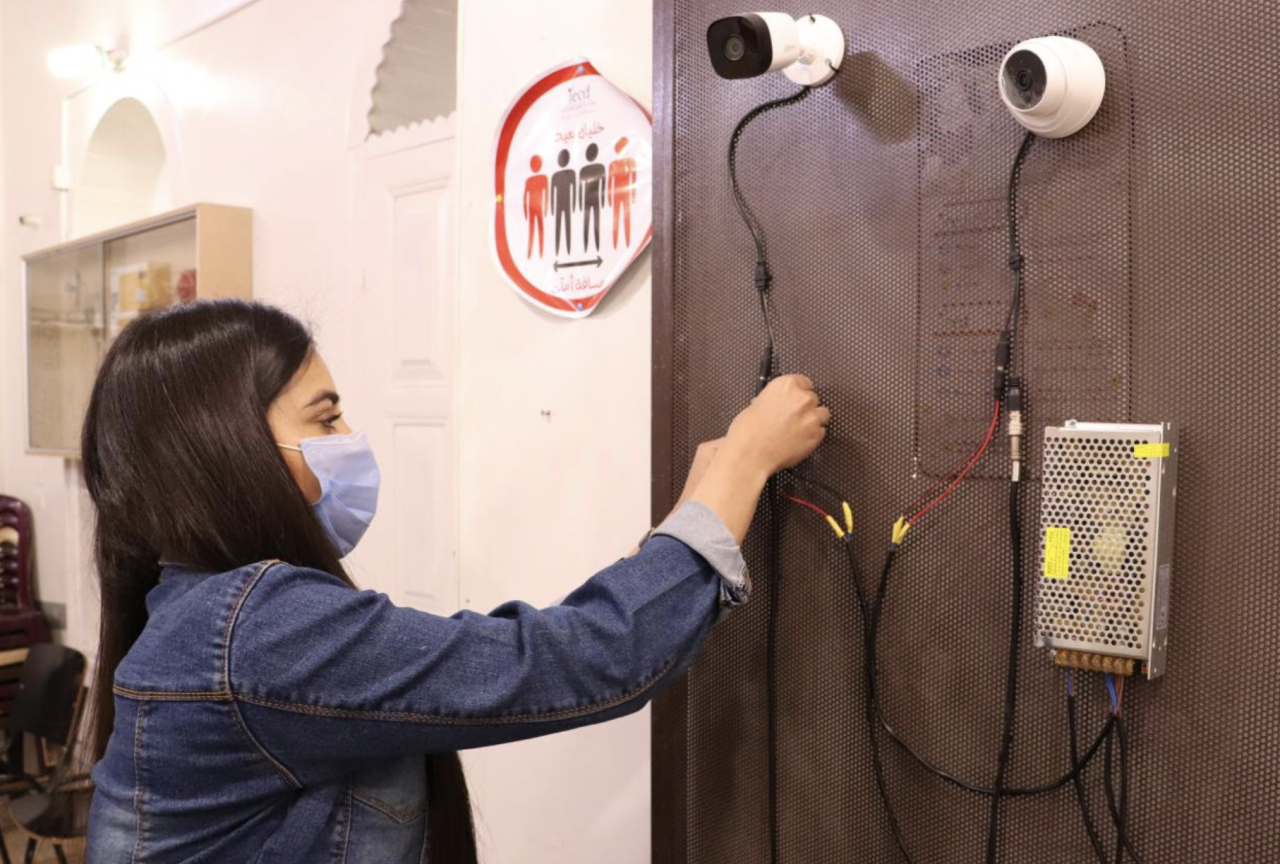 A girl in a blue face mask connects camera wires on a wall. 