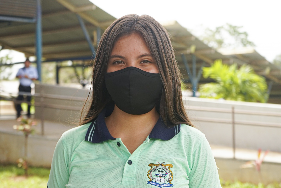 A girl in a school uniform and black face mask looks directly at the camera at her school. 