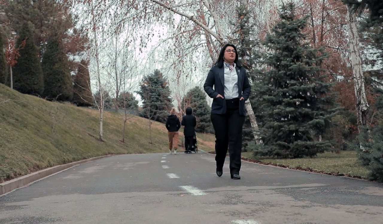 Aqida Mokhirova walks pensively on a park road, while the backs of two people, one of which pushes a baby stroller walk away from her.