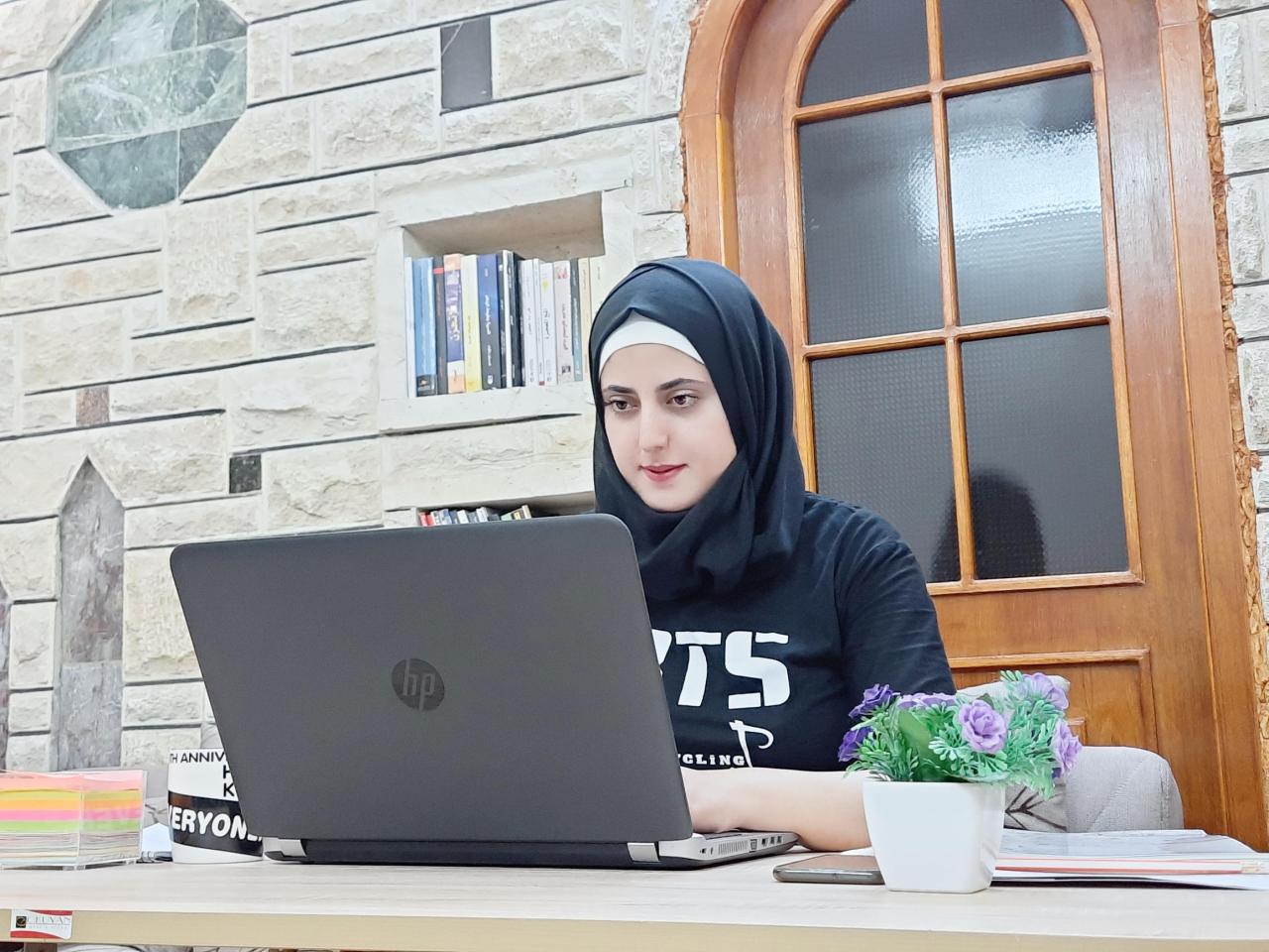A young woman sits in front of a lap top in front of a window.