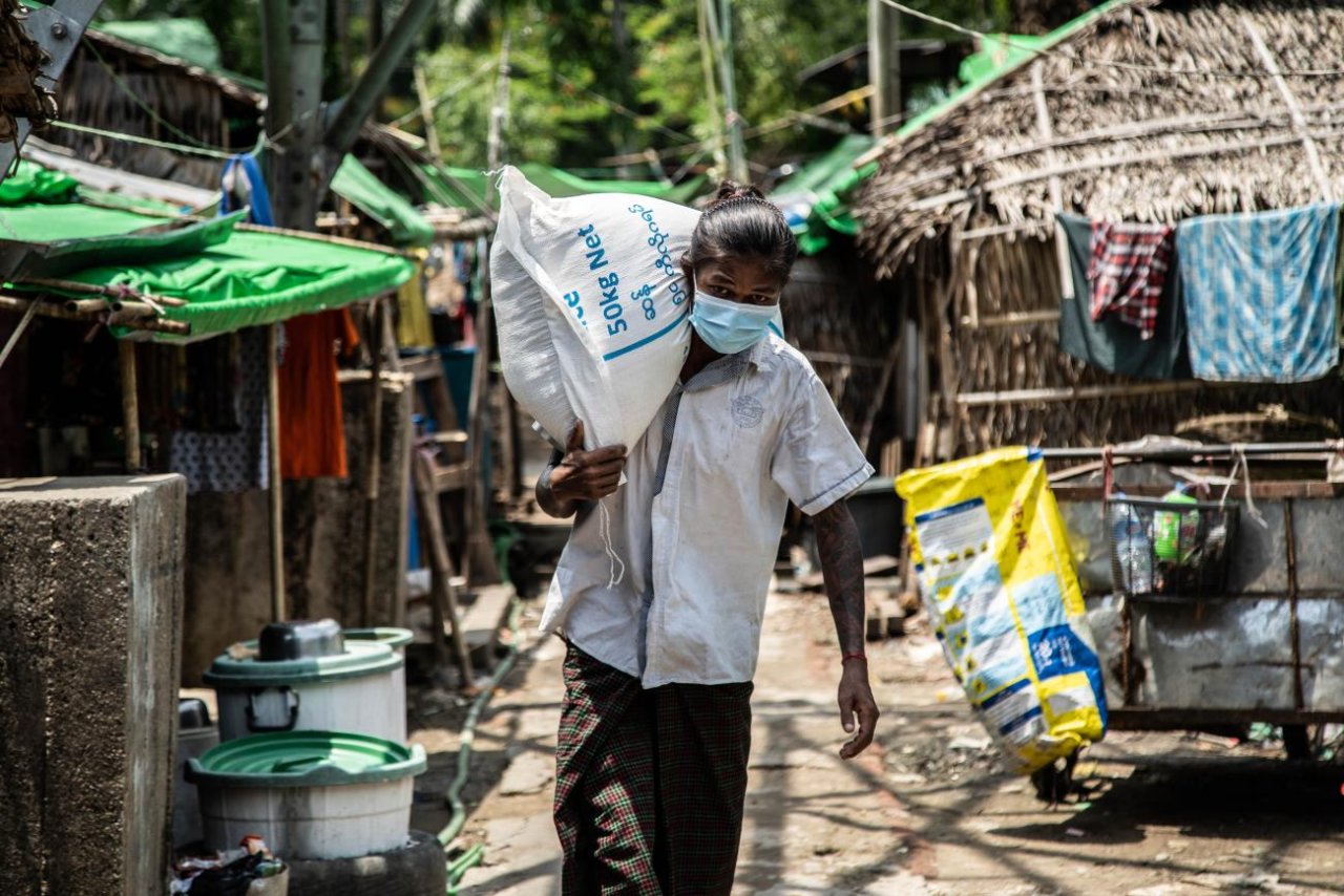 A person in a face mask carries a larg一个戴着面罩的人肩上扛着一个白色的大袋子，走在一条有很多人家的街道上。e white sack on their shoulder down a street with many homes. 