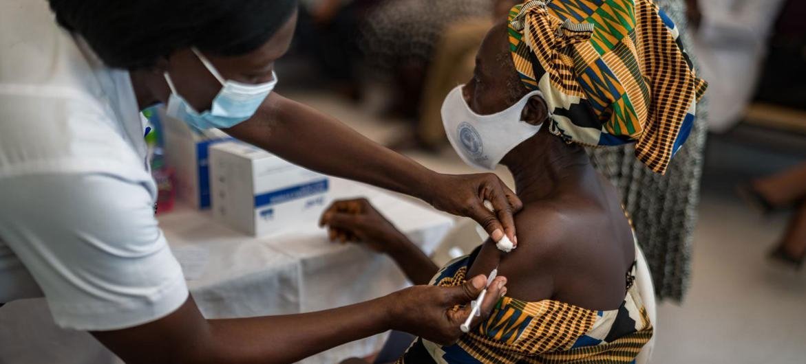 Une femme portant une blouse blanche et un masque de protection respiratoire administre un vaccin à une femme également masquée assise sur une chaise.
