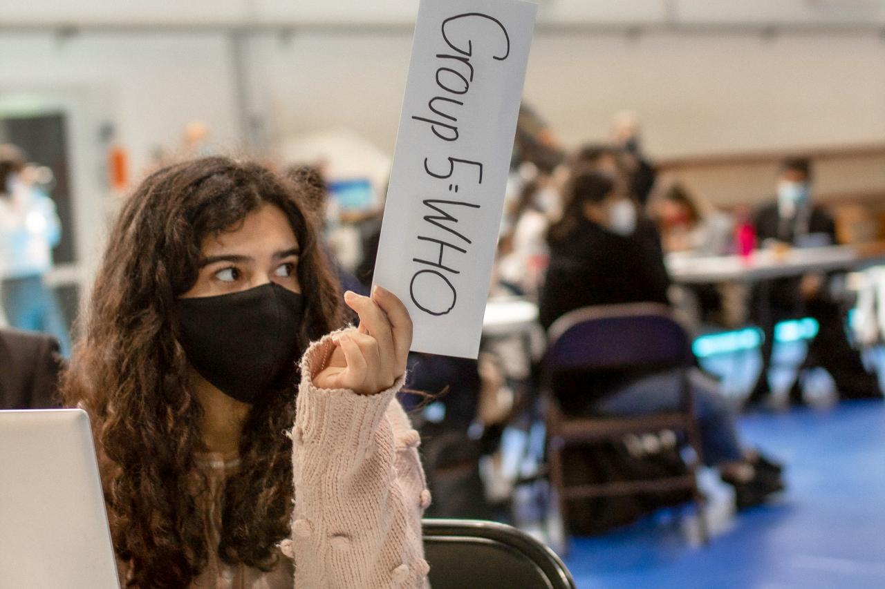 Une jeune fille aux cheveux châtains, longs et bouclés et portant un masque de protection respiratoire noir est assise dans une salle avec d’autres jeunes et brandit un écriteau de la main gauche. 