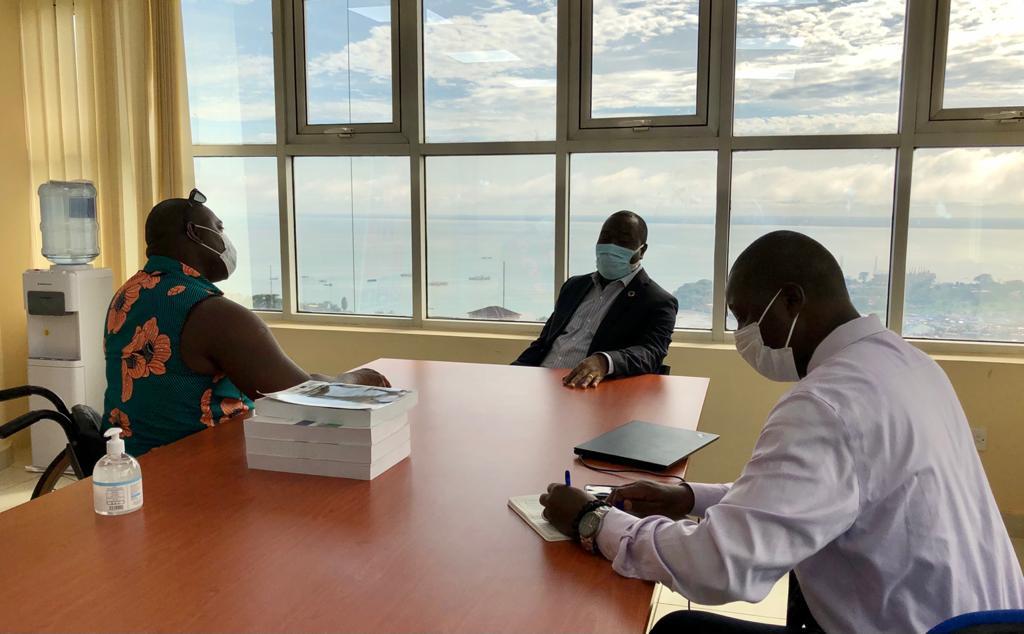 Three men sit around a table in face masks near a wall of windows. 