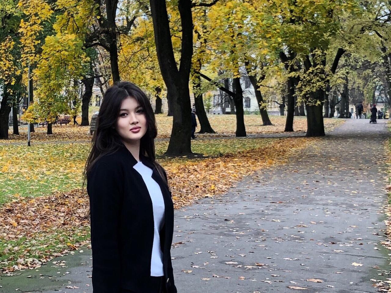 En Ouzbékistan, par une journée d’automne, une jeune fille aux longs cheveux noirs portant une veste noire se tient debout au milieu d’un sentier bordé d’arbres et de feuilles mortes en regardant la caméra de côté.