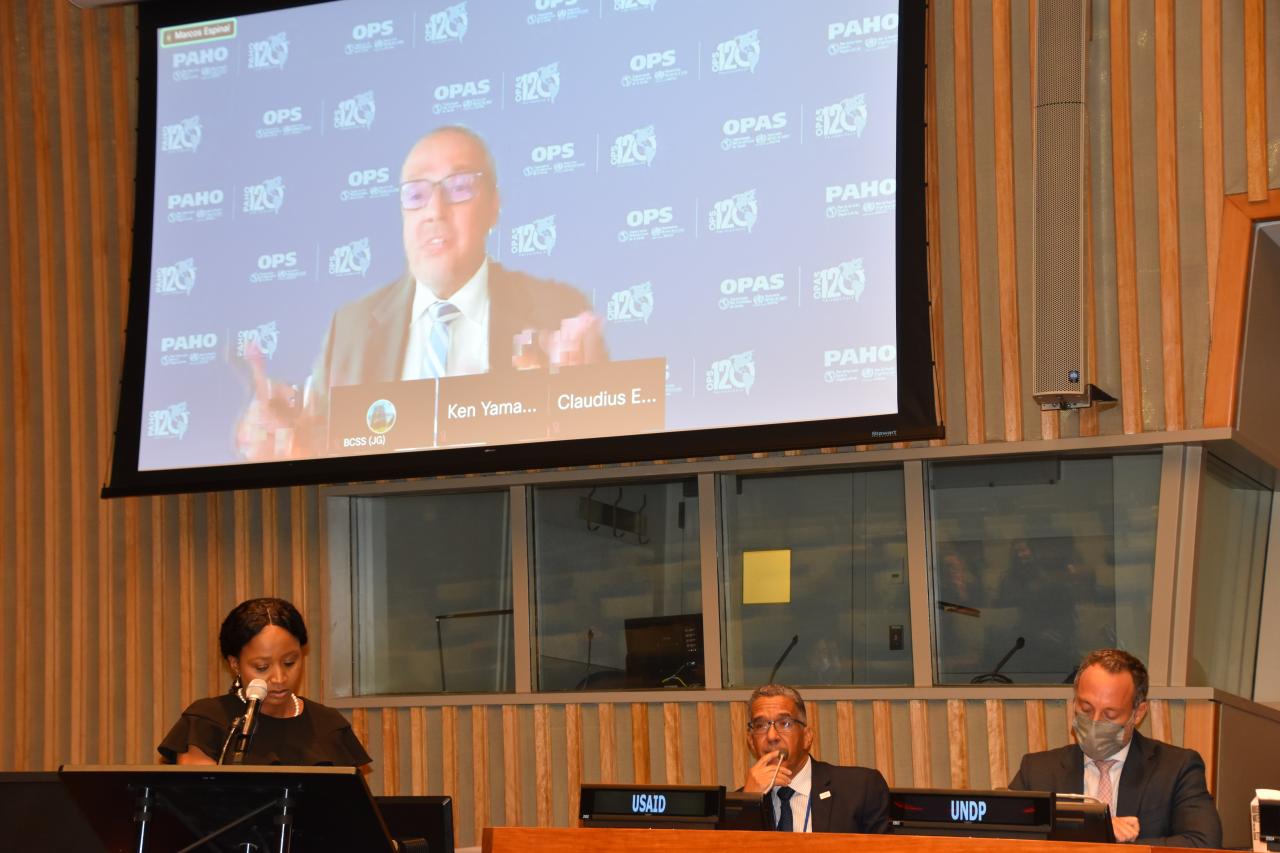 A woman speaks into a microphone, with men on a panel behind her, and a large screen showing an official speaking behind her as well.