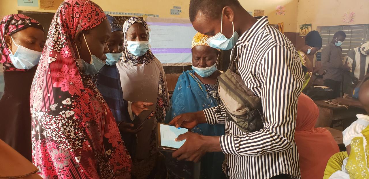 A young man wearing a face mask stands surrounded by several women, also masked. He holds a tablet and shows them how to use it. 